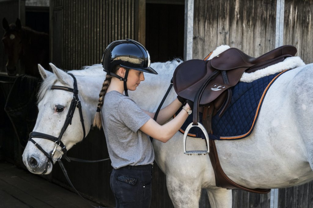 Teenage girl horserider with a grey horse outside astable, adjusting the girth and saddle.